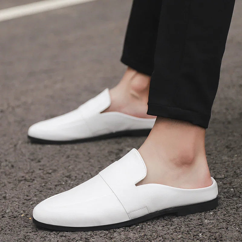 Men's white slip-on loafers with black soles worn with black pants on asphalt