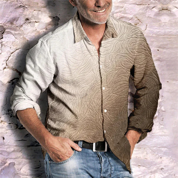 Man wearing a gradient beige and light brown patterned long-sleeve shirt with blue jeans against a textured white brick wall