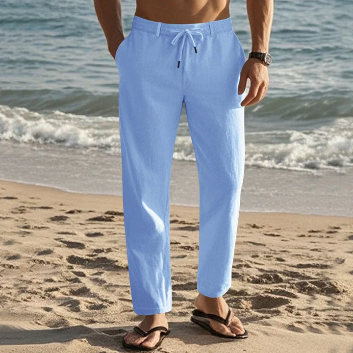 Man wearing light blue drawstring pants and black flip-flops standing on sandy beach near ocean
