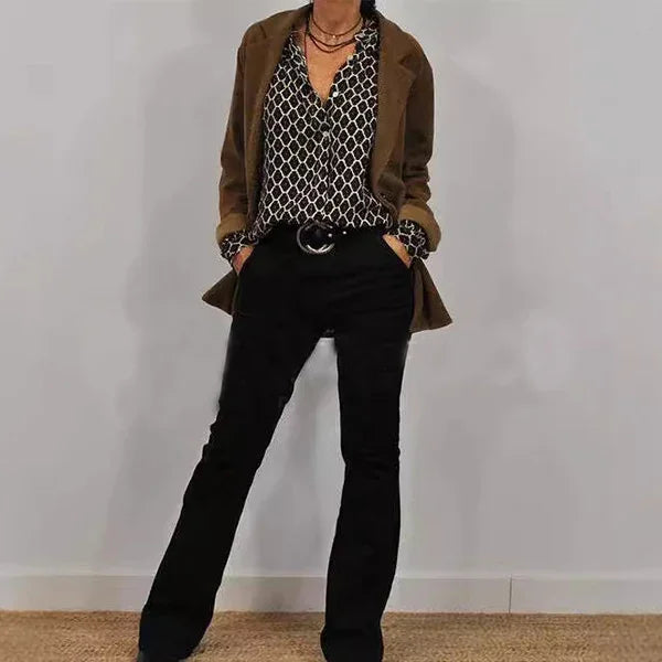 Woman wearing brown blazer, black patterned blouse, and black pants standing against gray wall
