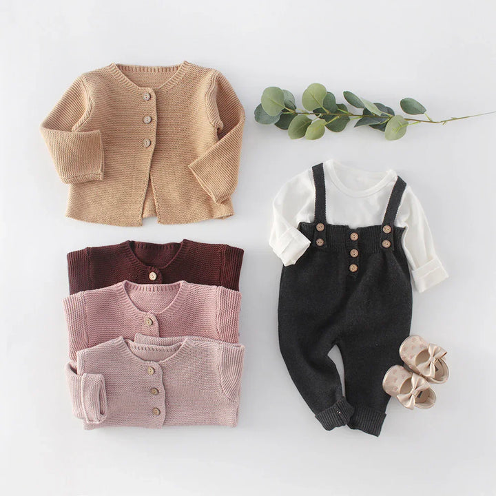 Set of knitted baby cardigans in beige, brown, mauve, and pink with a white long-sleeve top and black knit overalls with buttons, baby shoes, and green leaves