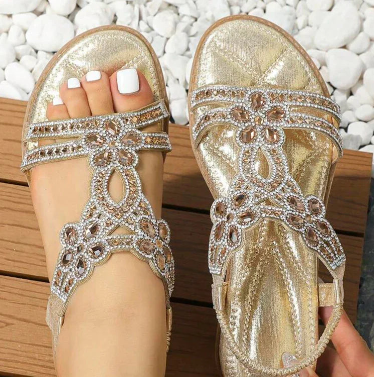 Women wearing gold glitter sandals with rhinestone floral design on wooden deck and white stones background