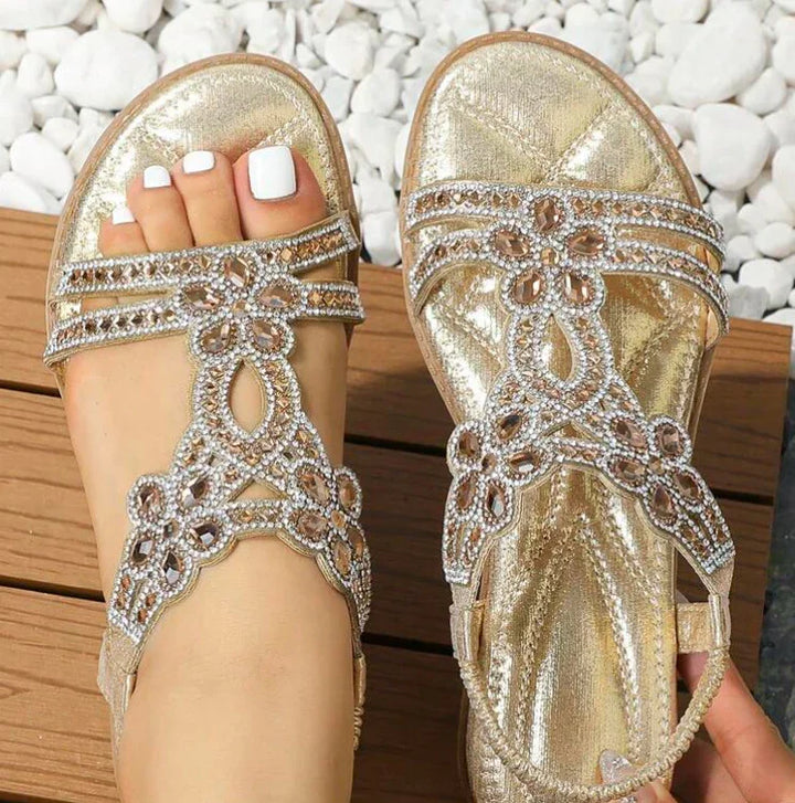 Women wearing gold glitter sandals with rhinestone floral design on wooden deck and white stones background