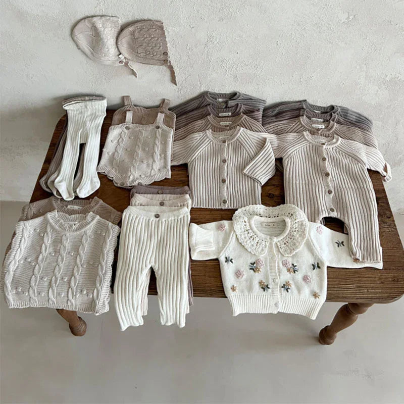 Neutral-toned knit baby clothes including sweaters, cardigans, pants, bonnets, and a floral embroidered sweater on a wooden table