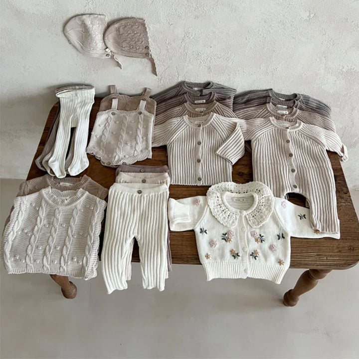 Neutral-toned knit baby clothes including sweaters, cardigans, pants, bonnets, and a floral embroidered sweater on a wooden table