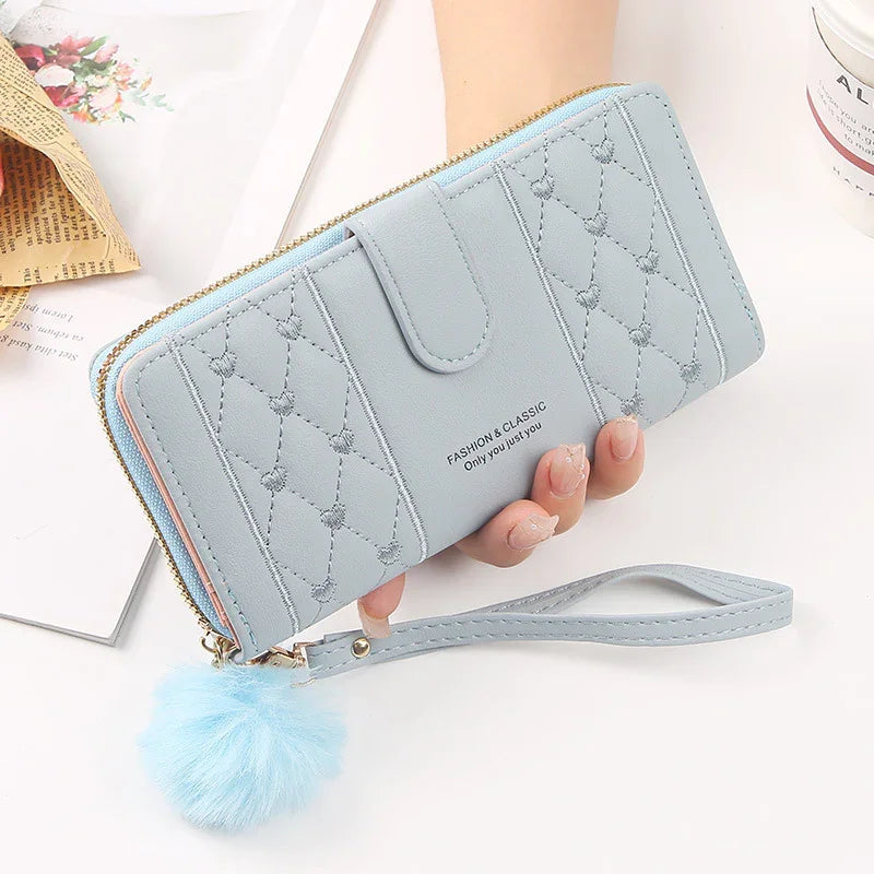 Light blue quilted wallet with heart patterns, zipper closure, wrist strap, and fluffy pom-pom accessory