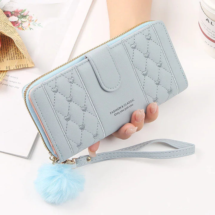 Light blue quilted wallet with heart patterns, zipper closure, wrist strap, and fluffy pom-pom accessory