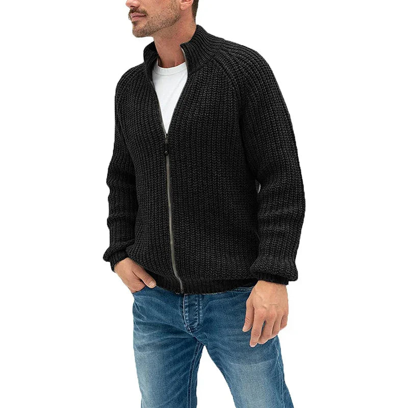 Man wearing black chunky knit zip-up sweater paired with blue jeans on white background