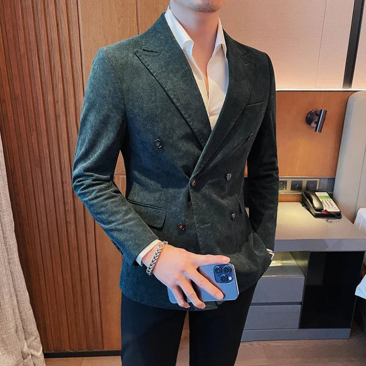Man wearing dark green double-breasted velvet blazer with white shirt, holding blue smartphone indoors
