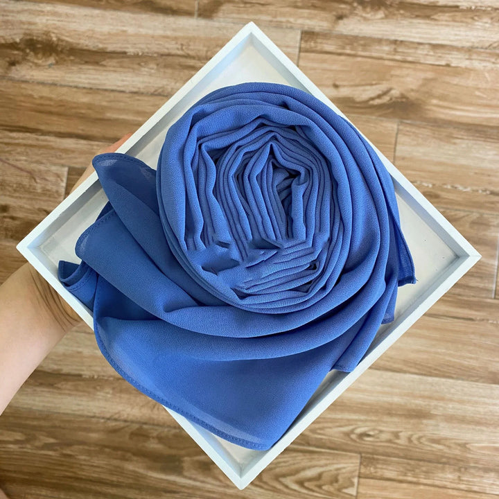 Hand holding white square tray with neatly folded blue fabric scarf on wooden floor background
