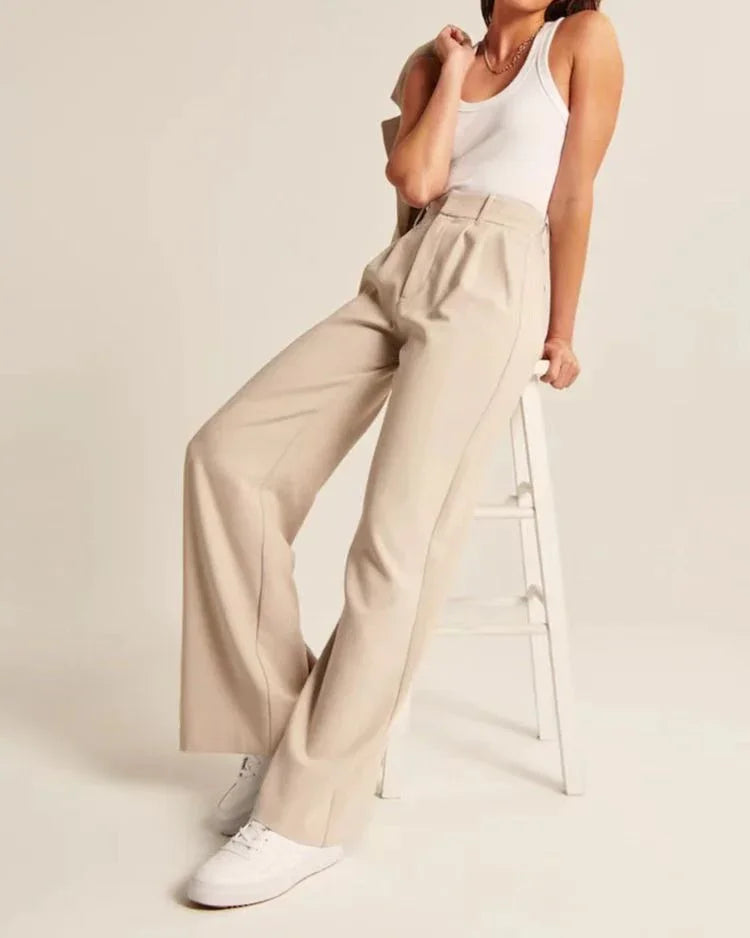 Woman wearing beige wide-leg pants and white tank top seated on white stool in studio