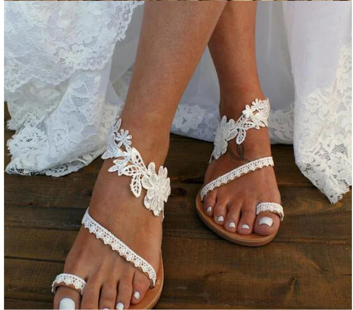 Close-up of bride's feet wearing white lace floral wedding sandals on wooden floor with lace dress hem