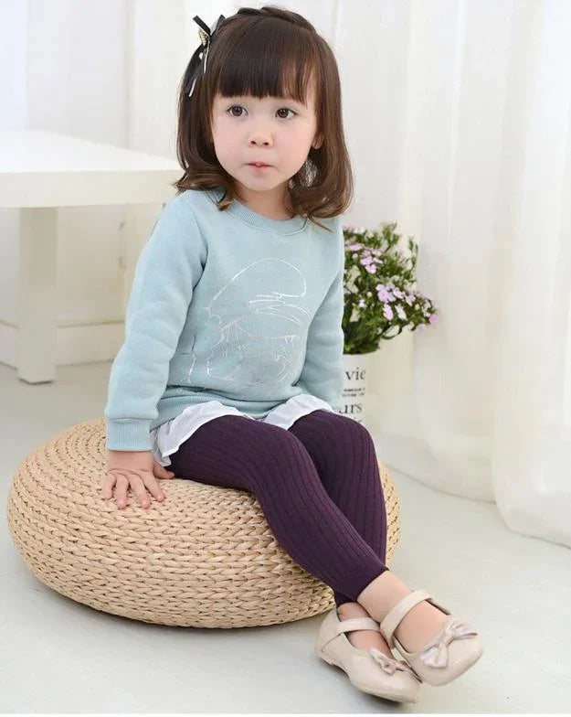 Little girl sitting on a woven round cushion wearing a blue sweater, purple leggings, and beige shoes