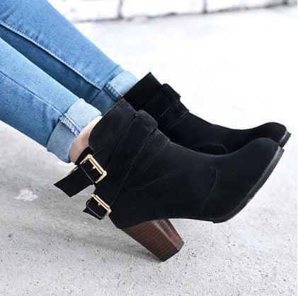 Black suede ankle boots with double gold buckles and chunky wooden heels worn with rolled-up blue jeans on a concrete floor