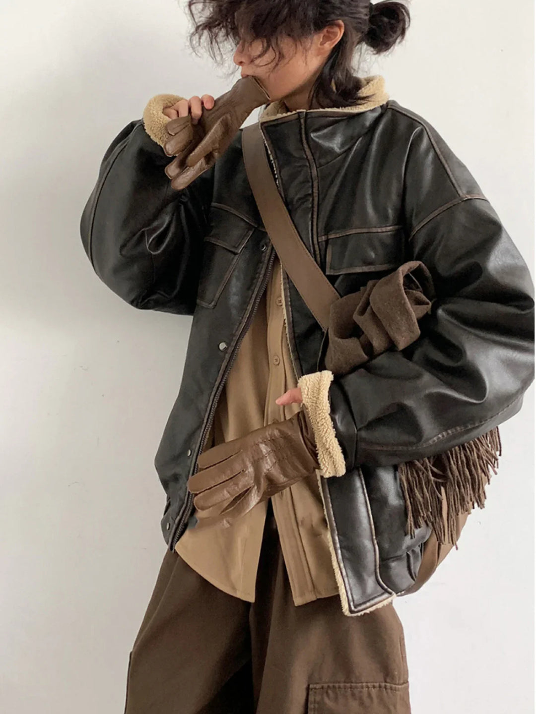 Person wearing black shearling leather jacket, brown gloves, beige shirt, and carrying brown fringed bag