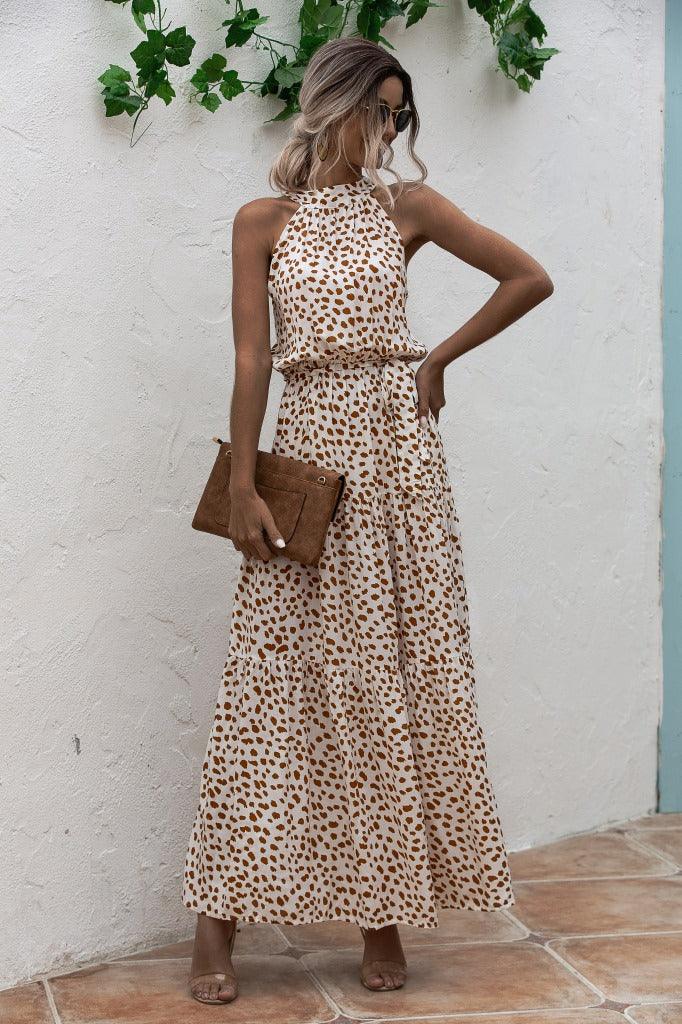 Woman wearing sleeveless beige maxi dress with brown spots holding a brown clutch bag against white textured wall with green leaves