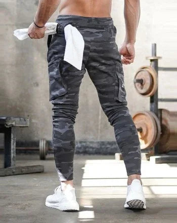 Man wearing black camo jogger pants and white sneakers in gym setting holding towel