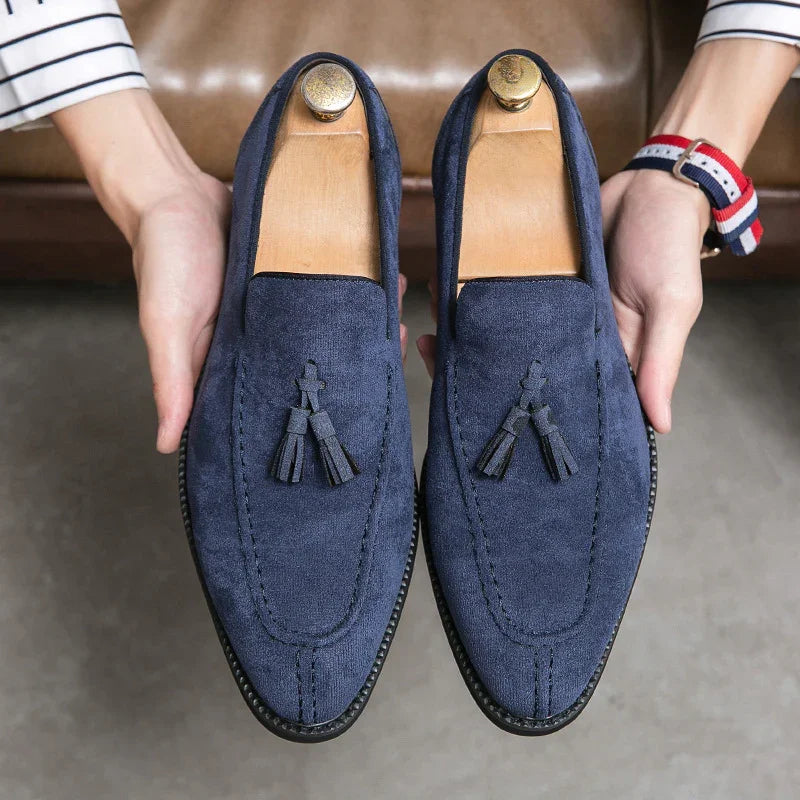 Hands holding navy blue suede tassel loafers with wooden shoe trees, leather soles, and stitched detailing