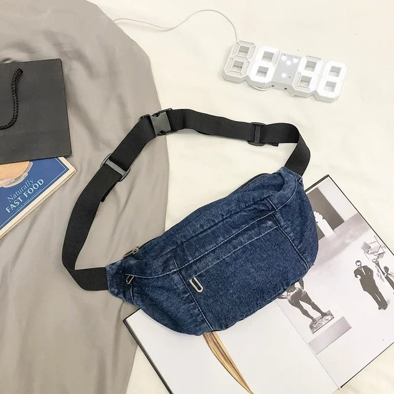 Denim blue adjustable waist bag with black strap on beige surface next to open magazine and digital clock