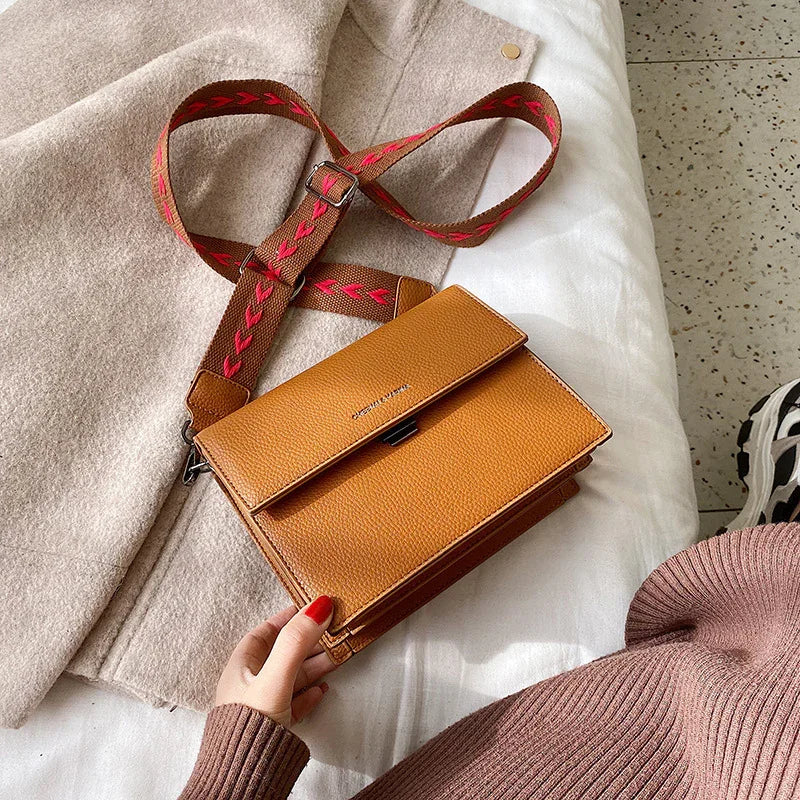 Tan leather crossbody bag with brown and red adjustable strap on beige coat and white bed