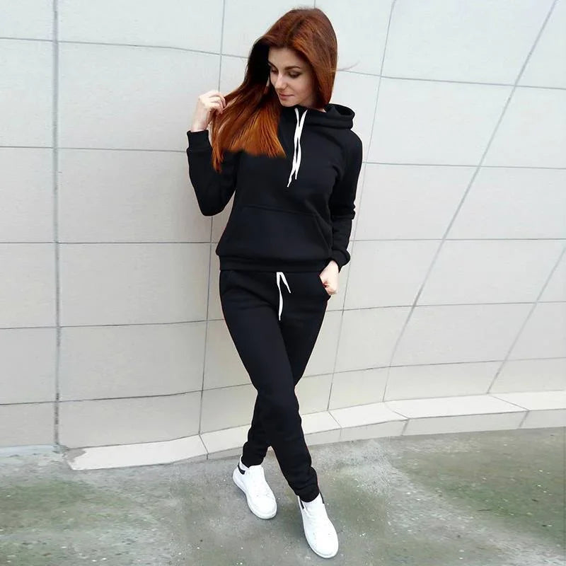 Woman wearing black hoodie and joggers with white drawstrings and white sneakers against tiled wall