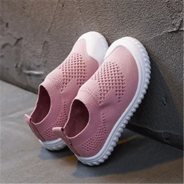 Pink knitted slip-on shoes with white soles resting against a textured wall