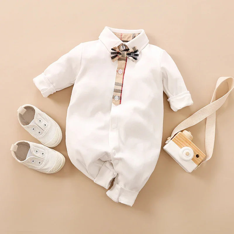 White baby romper with plaid bow tie and button detail, paired with white baby shoes and wooden toy camera