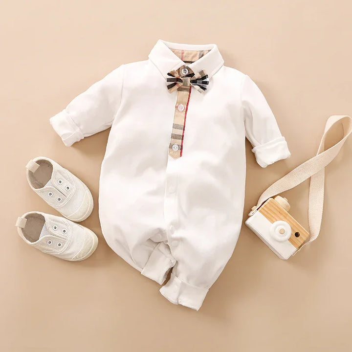 White baby romper with plaid bow tie and button detail, paired with white baby shoes and wooden toy camera