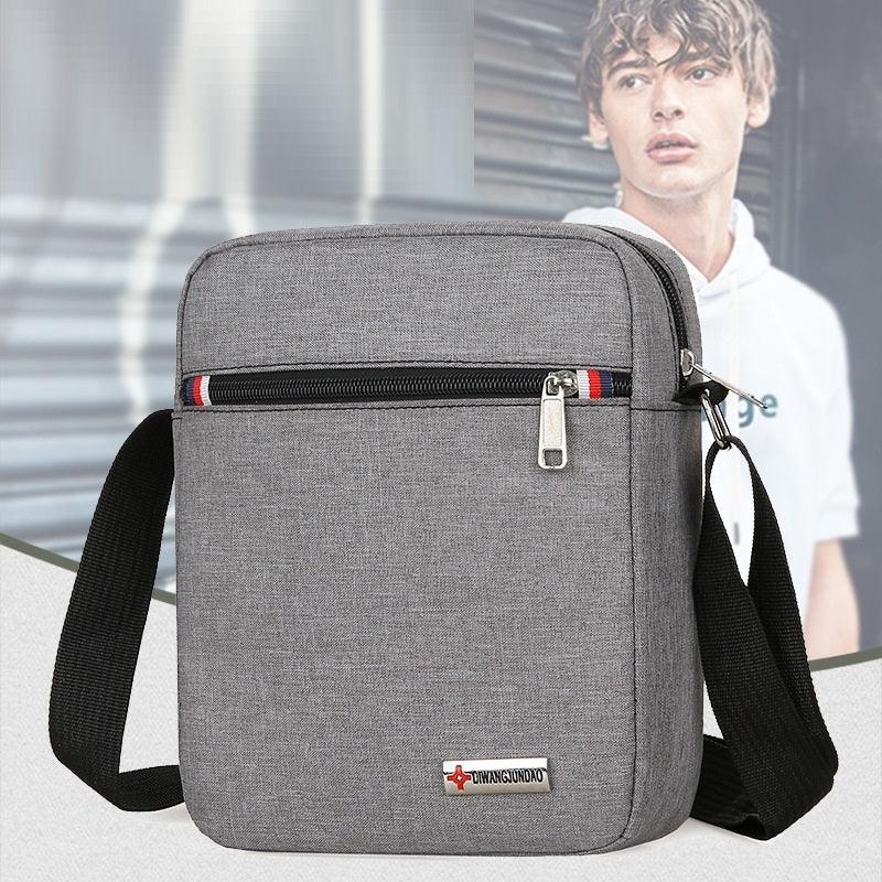 Gray crossbody bag with black adjustable strap and front zipper pocket, featuring a small logo tag