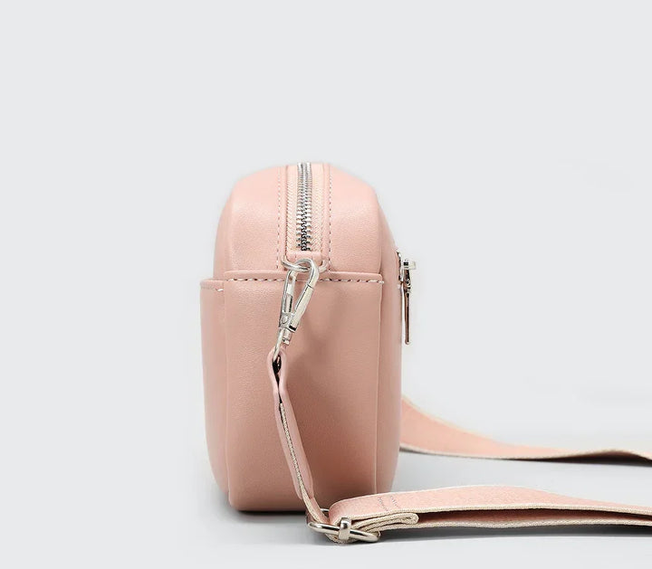 Side view of a small pink crossbody bag with silver zipper and adjustable strap on grey background
