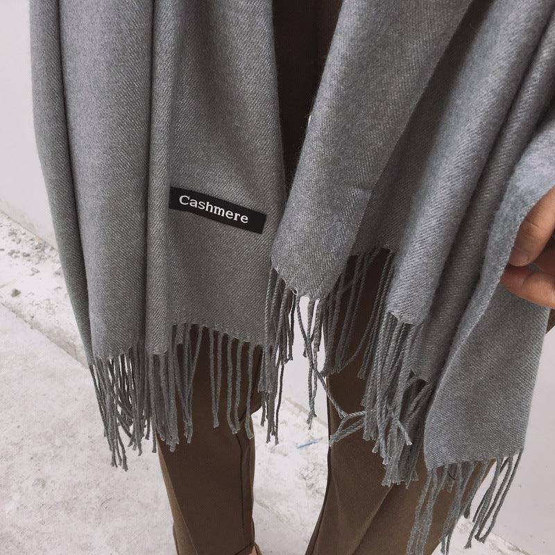Soft gray cashmere scarf with fringed edges draped over brown pants on a concrete floor