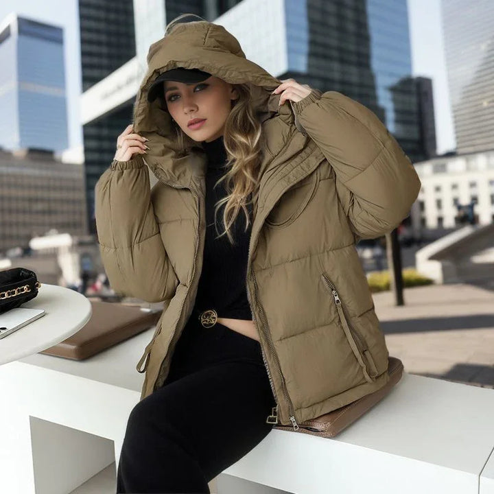 Woman in a tan puffer jacket with hood, black top, and pants sitting outdoors in an urban setting