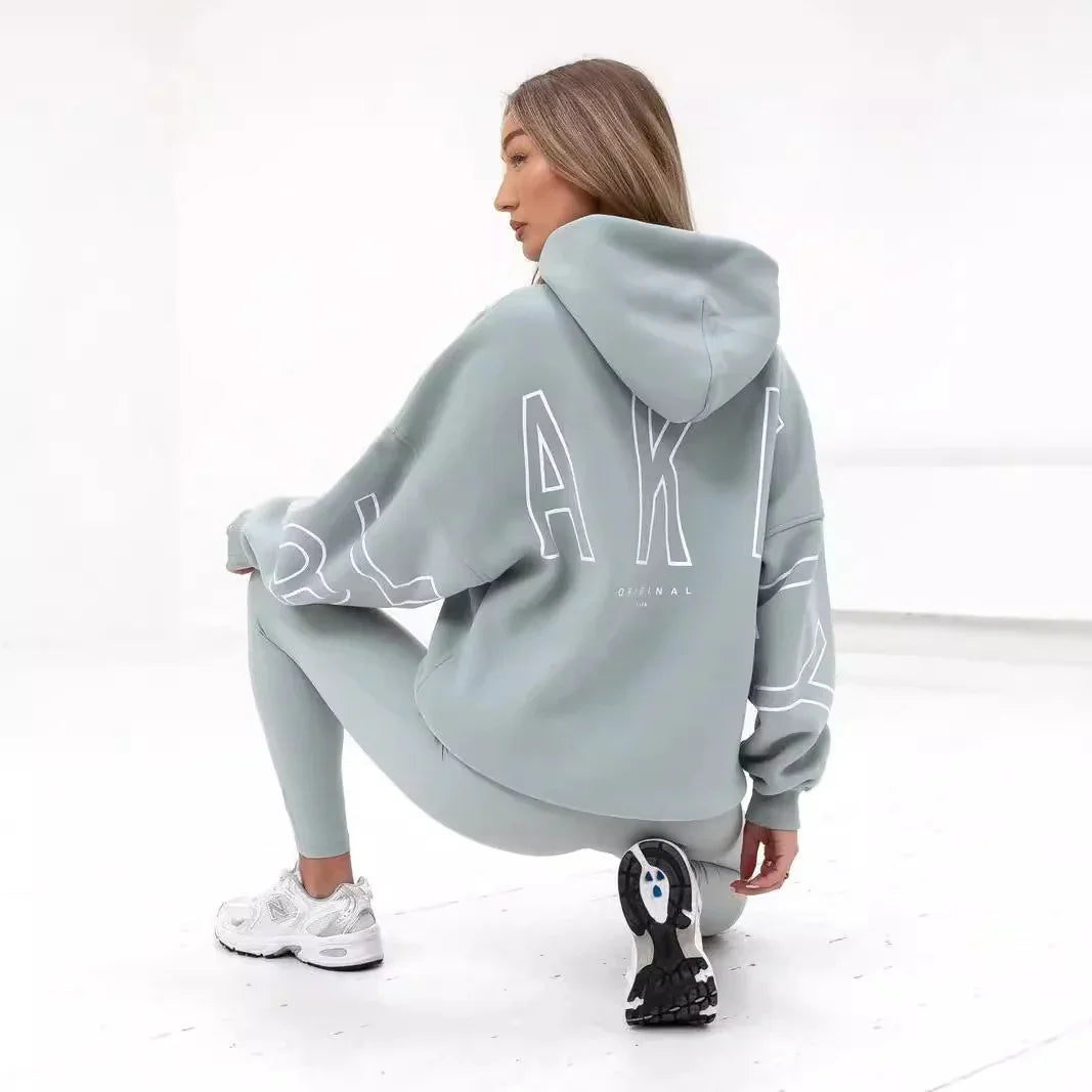 Woman squatting in pastel green hoodie with large white lettering and matching leggings, wearing white New Balance sneakers on white floor