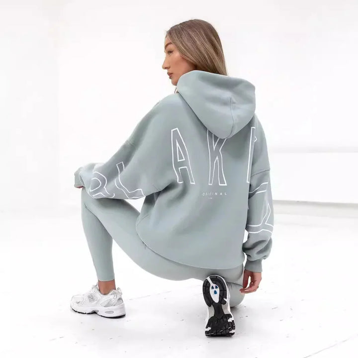 Woman squatting in pastel green hoodie with large white lettering and matching leggings, wearing white New Balance sneakers on white floor