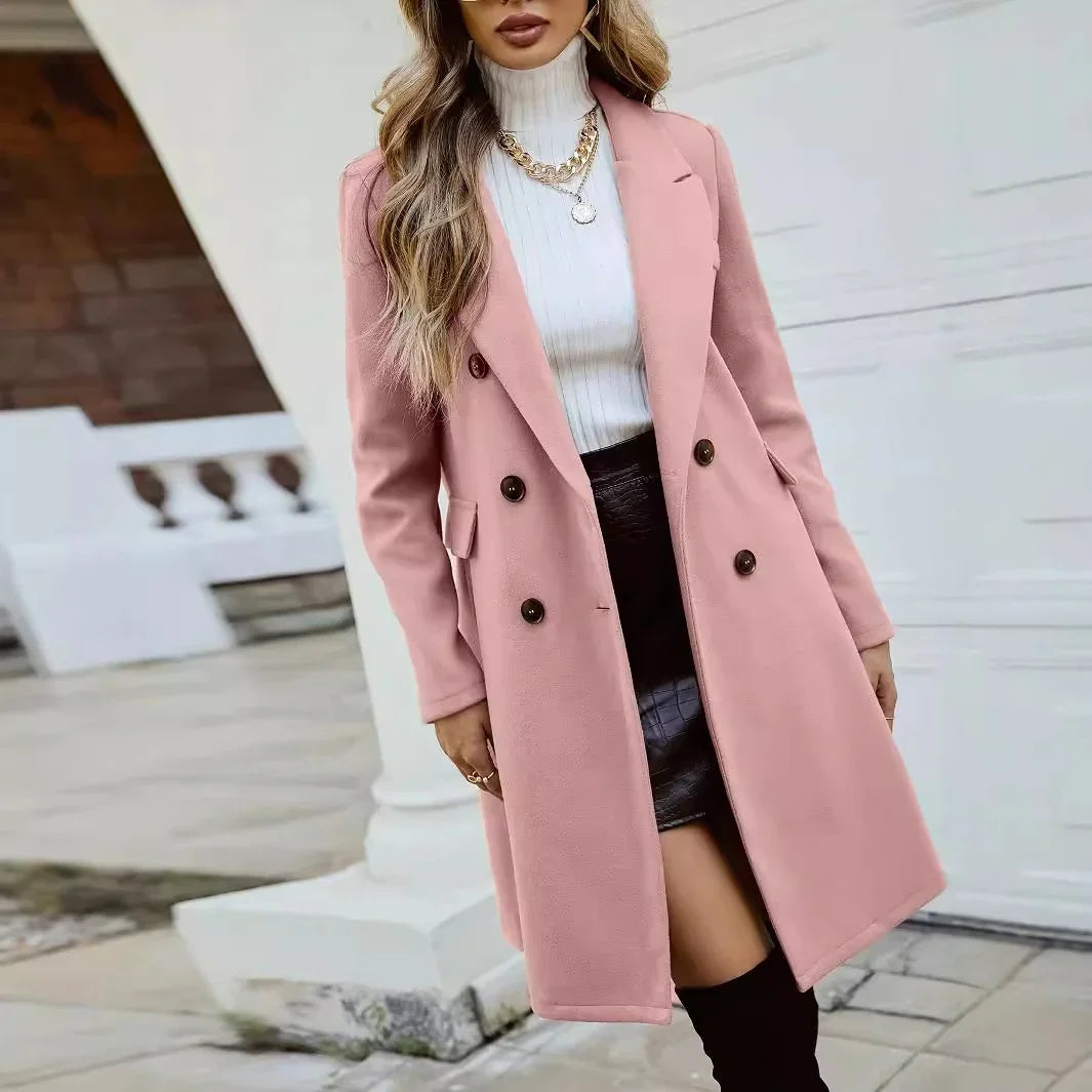 Woman wearing a pink double-breasted coat, white turtleneck sweater, black skirt, and gold necklaces in outdoor setting