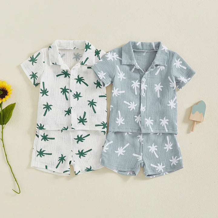Two toddler summer outfits with palm tree prints, one white with green and one light blue with white, laid flat