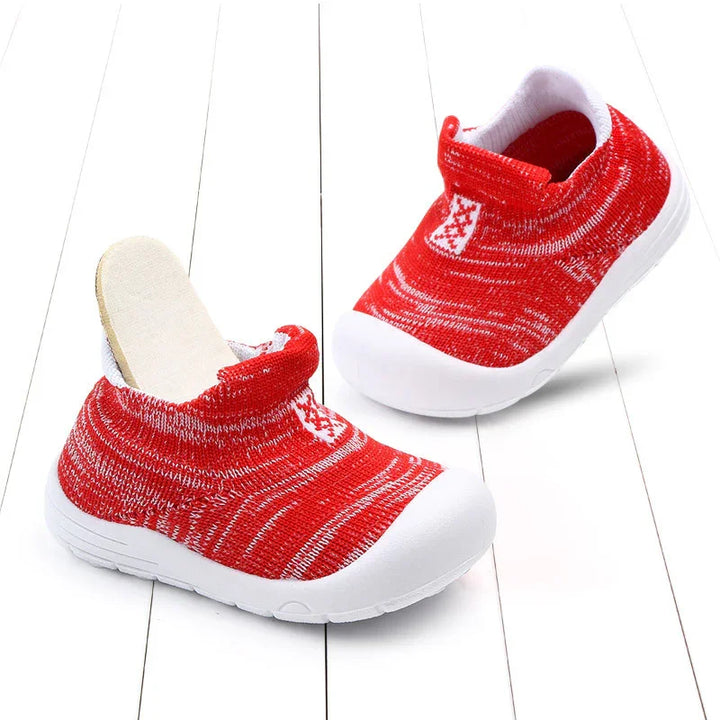 Red and white toddler slip-on shoes with flexible knit fabric and white rubber soles on white wooden floor