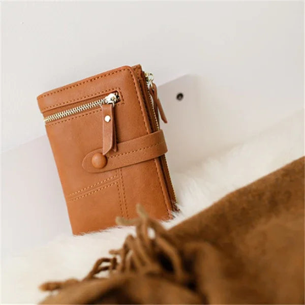 Brown leather wallet with zipper and button strap on a soft white surface, with blurred fabric in foreground
