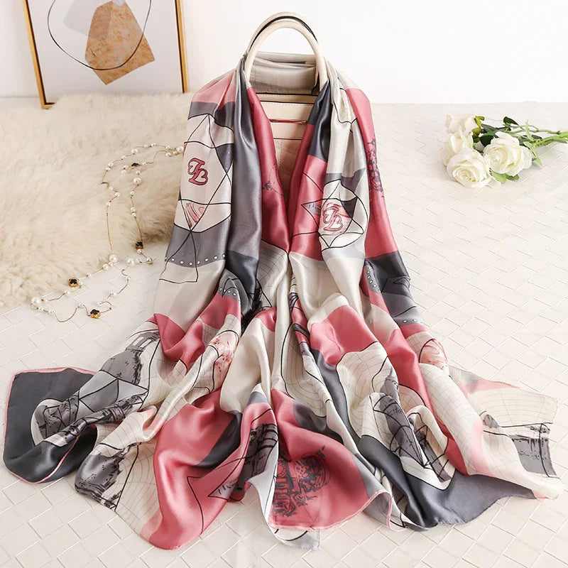 Soft silk scarf with pink, gray, and white geometric patterns draped over beige handbag, white roses and pearl necklace nearby