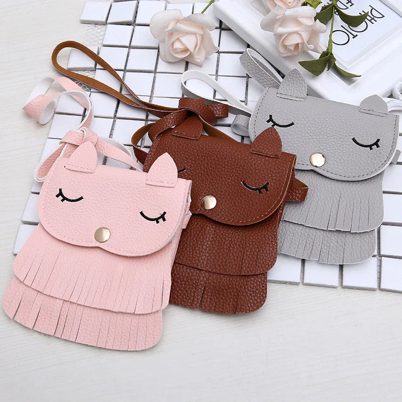 Three leather crossbody bags in pink, brown, and gray with cat ear designs and eyelash details on white tiled surface
