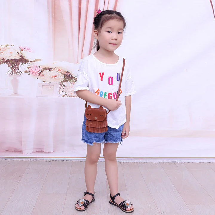 Young girl in white graphic t-shirt, denim shorts, black sandals, and brown cat-shaped crossbody bag
