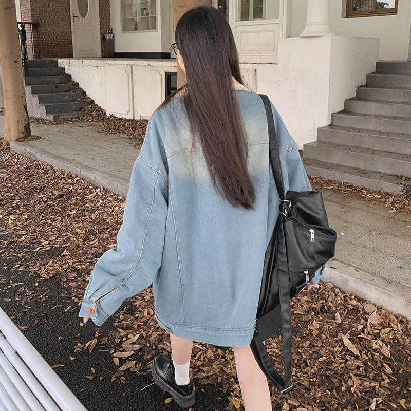 Woman wearing oversized light blue denim jacket and black shoes walking on leaf-covered sidewalk carrying black backpack