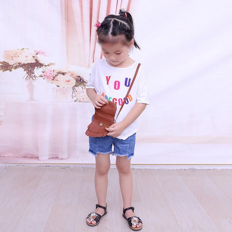 Young girl wearing colorful t-shirt, denim shorts, black sandals, holding brown fringed leather purse indoors