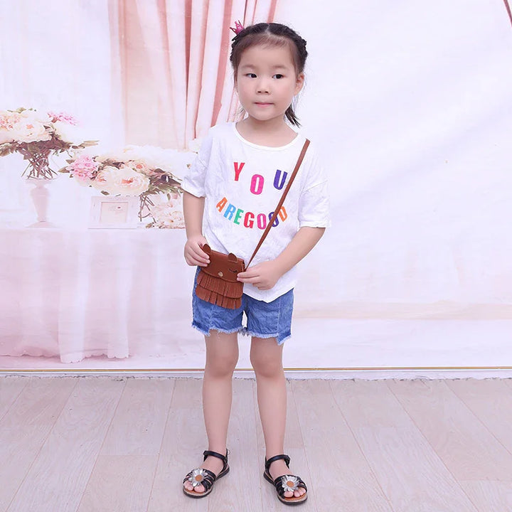 Young girl wearing white t-shirt with colorful text, denim shorts, brown crossbody bag, and black sandals