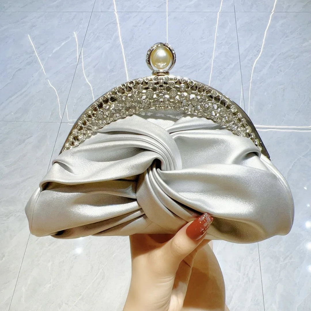 Hand holding a silver satin evening clutch with pearl and rhinestone clasp on marble floor