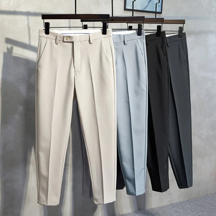 Four pairs of formal trousers in beige, light blue, black, and gray hanging on wooden hangers against a gray wall