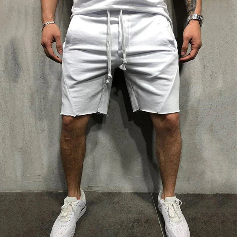 Man wearing casual white drawstring shorts and white sneakers standing against gray wall