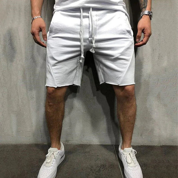 Man wearing casual white drawstring shorts and white sneakers standing against gray wall