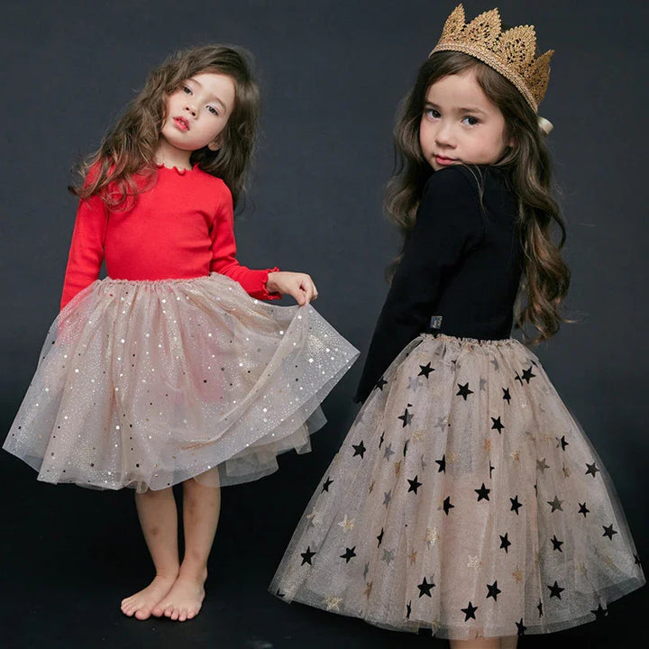 Two girls in glittery tulle skirts, one in red top, the other in black top with gold crown on dark background