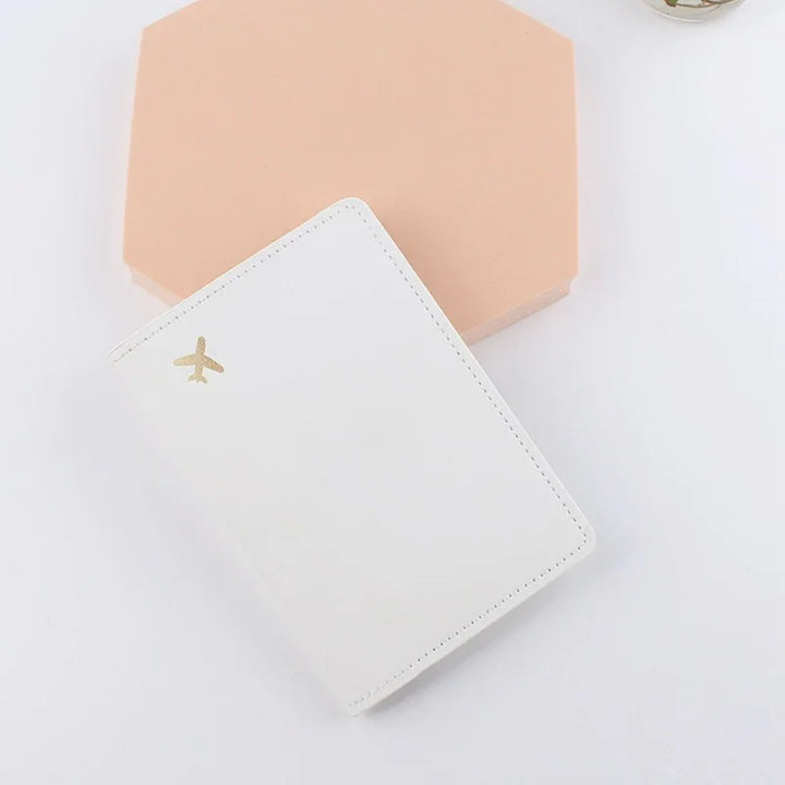 White leather passport holder with gold airplane icon on a light pink hexagonal surface